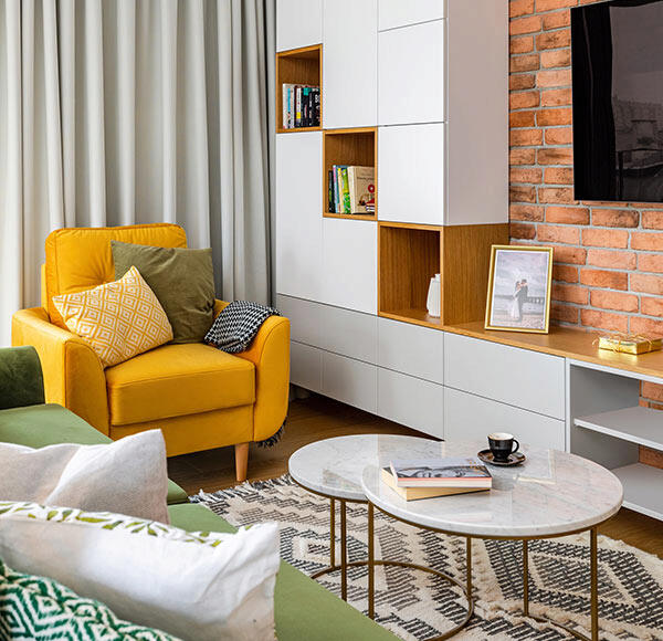 Nowoczesny salon z żółtym fotelem, ceglaną ścianą i minimalistyczną zabudową meblową w bieli z drewnianymi akcentami.